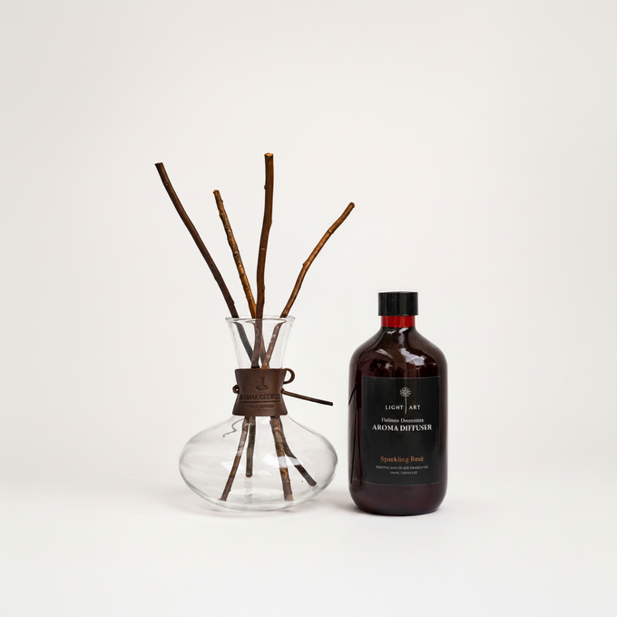 A bottle of Light Art Sparkling Rose aroma diffuser next to a clear glass vase with brown reed sticks, on a clean white background.