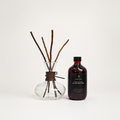 A bottle of Light Art Sparkling Rose aroma diffuser next to a clear glass vase with brown reed sticks, on a clean white background.