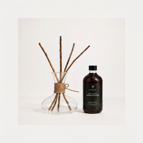 A reed diffuser set on a clean white background, featuring a clear glass vase with several natural brown reeds tied in the neck with a tan paper band, next to a dark amber bottle of Light Art 'Spiked Cedar' aroma diffuser liquid.