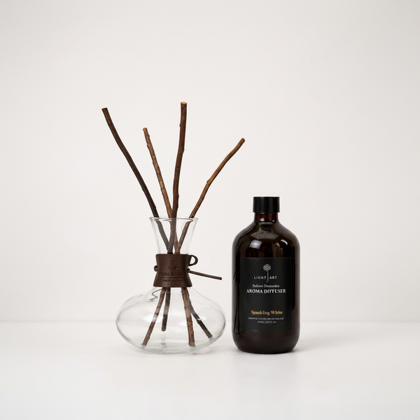 A reed diffuser set on a white background, featuring a clear glass bottle with a dark brown label reading 'Light Art' and 'Aroma Diffuser', next to a clear glass vase with brown reeds and a dark brown leather tie.