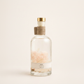 A clear glass perfume bottle with a gold cap and a beige paper neck label, containing pale pink rose petals suspended in clear liquid. The bottle is centered against a solid, warm off-white background.