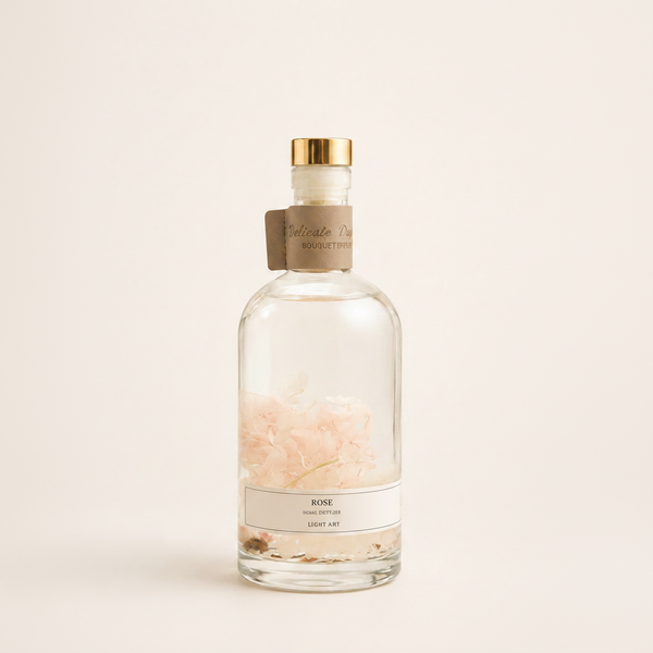 A clear glass perfume bottle with a gold cap and a beige paper neck label, containing pale pink rose petals suspended in clear liquid. The bottle is centered against a solid, warm off-white background.