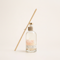 A clear glass reed diffuser bottle with a white label and gold cap, filled with pale pink wax flakes, with two natural wood reeds leaning against it on a warm off-white background.