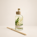 A clear glass bottle of reed diffuser with a gold cap and a kraft paper neck label, filled with pale liquid and containing dried green and cream botanical elements. A pair of natural bamboo reeds lies on the surface in front, against a clean off-white background.