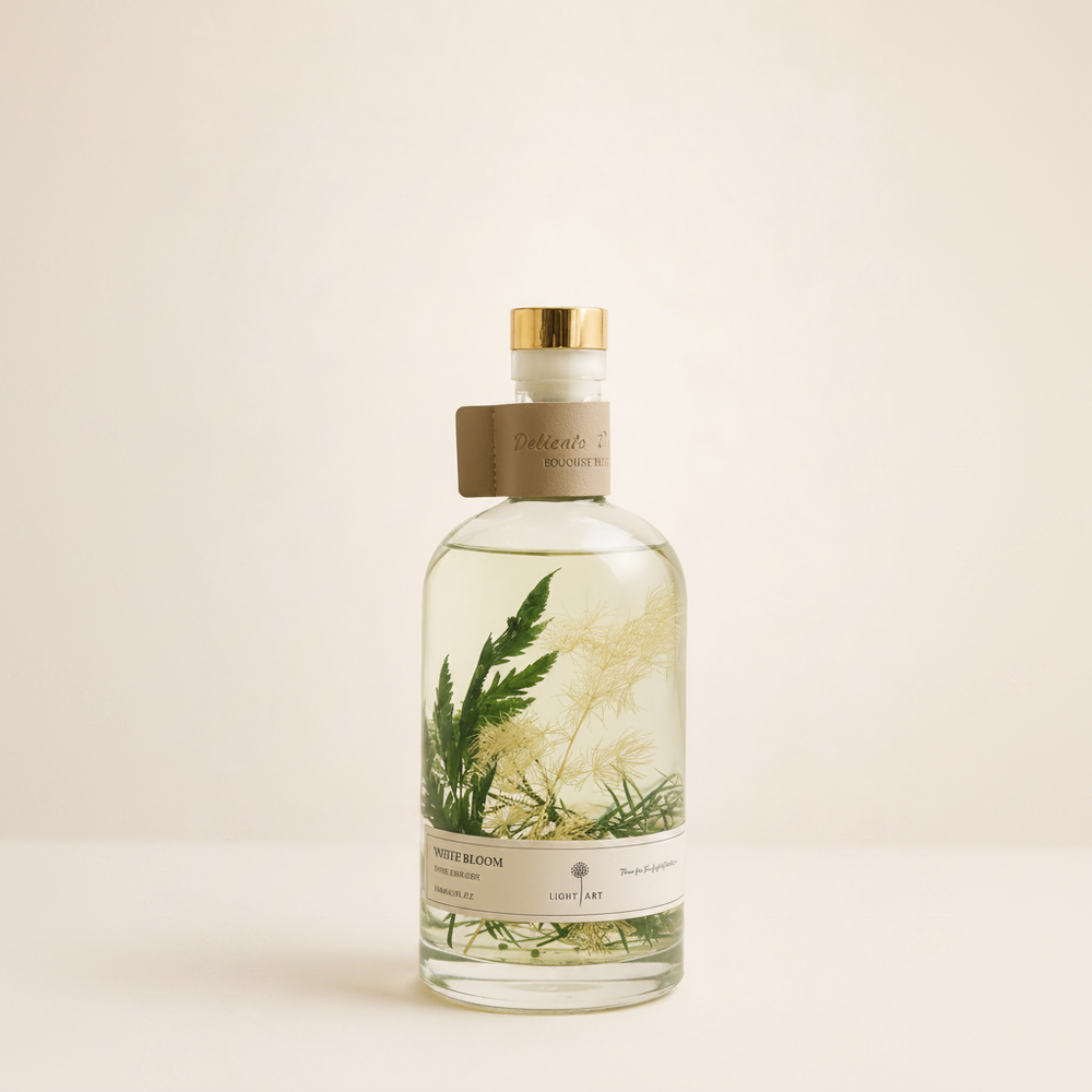 A clear glass bottle of botanical-infused liquid with a gold cap and a brown paper collar, featuring pressed green and beige botanical elements inside. The bottle is set against a solid, warm off-white background.