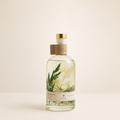 A clear glass bottle of botanical-infused liquid with a gold cap and a brown paper collar, featuring pressed green and beige botanical elements inside. The bottle is set against a solid, warm off-white background.