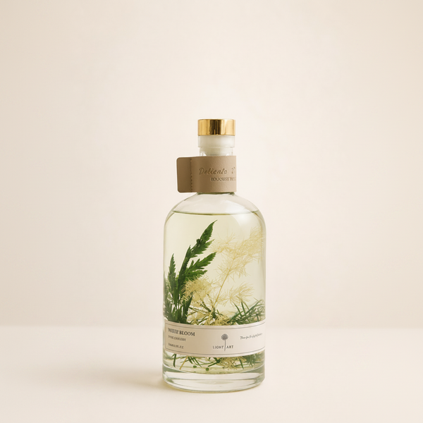 A clear glass bottle of botanical-infused liquid with a gold cap and a brown paper collar, featuring pressed green and beige botanical elements inside. The bottle is set against a solid, warm off-white background.