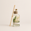 A clear glass reed diffuser bottle with a gold cap, a brown paper collar, and a white label reading 'White Bloom', filled with pale liquid and botanicals. Two natural wood reeds lean against the bottle on a warm off-white background.